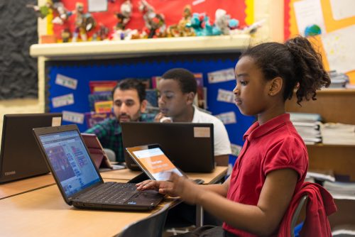 A boy and a girl aged around 9-11 years sit at a desk in a classroom, focussed on the activity they are doing using laptops. From the girl's screen we can see that she is programming in Scratch. A man sits beside the boy, helping him with his work.